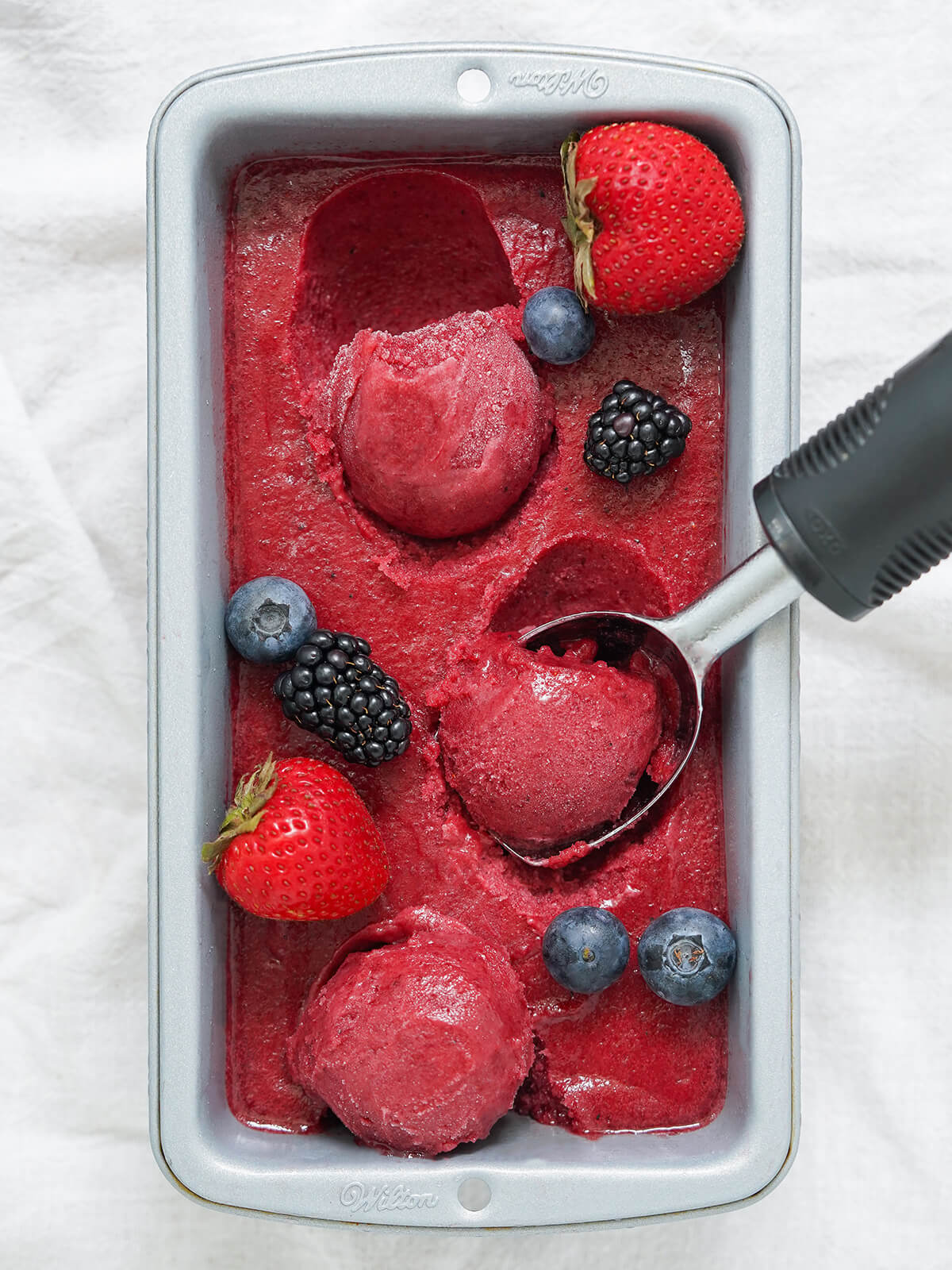 overhead pic of container of mixed berry sorbet with scoops formed sitting on top, one in the ice cream scoop.
