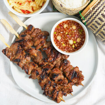 three moo ping Thai pork skewers on plate with dish of dipping sauce to side.