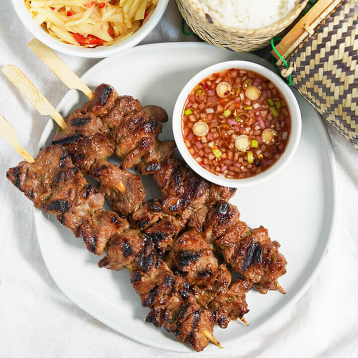 three moo ping Thai pork skewers on plate with dish of dipping sauce to side.