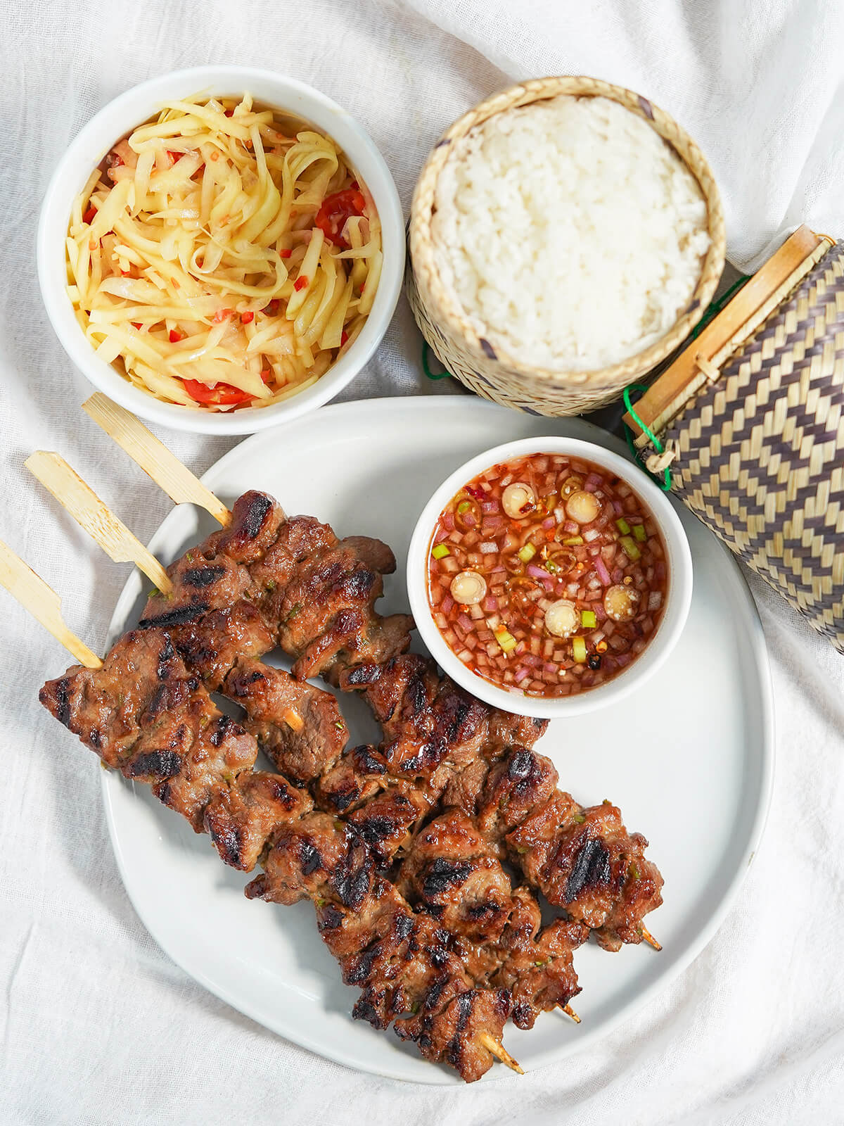 Moo ping are delicious Thai pork skewers, with a sweet, salty, umami marinade that packs in flavor and gives a caramelized edge. Delicious as a snack or a main with simple sides. overhead view of plate of moo ping, Thai pork skewers, with small bowl of dipping sauce on one side and bowls of papaya salad and sticky rice above.