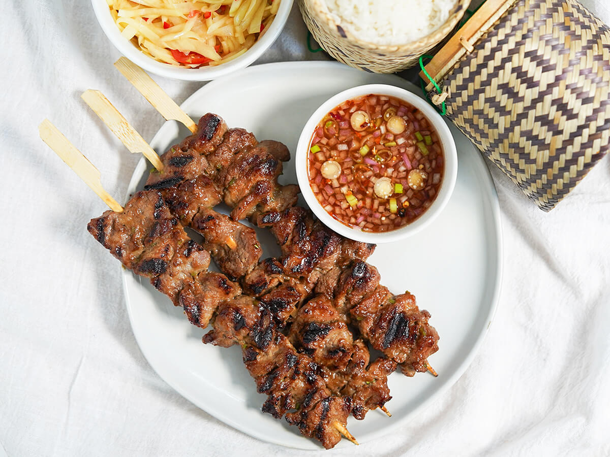 moo ping Thai pork skewers overhead view of plate of moo ping Thai pork skewers with dipping sauce and edge of dishes of papaya salad and rice showing at top.