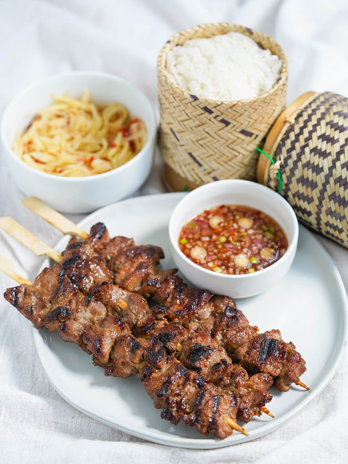 Moo ping are delicious Thai pork skewers, with a sweet, salty, umami marinade that packs in flavor and gives a caramelized edge. Delicious as a snack or a main with simple sides. side view of plate of moo ping Thai pork skewers with dipping sauce, bowl of papaya salad and sticky rice behind.