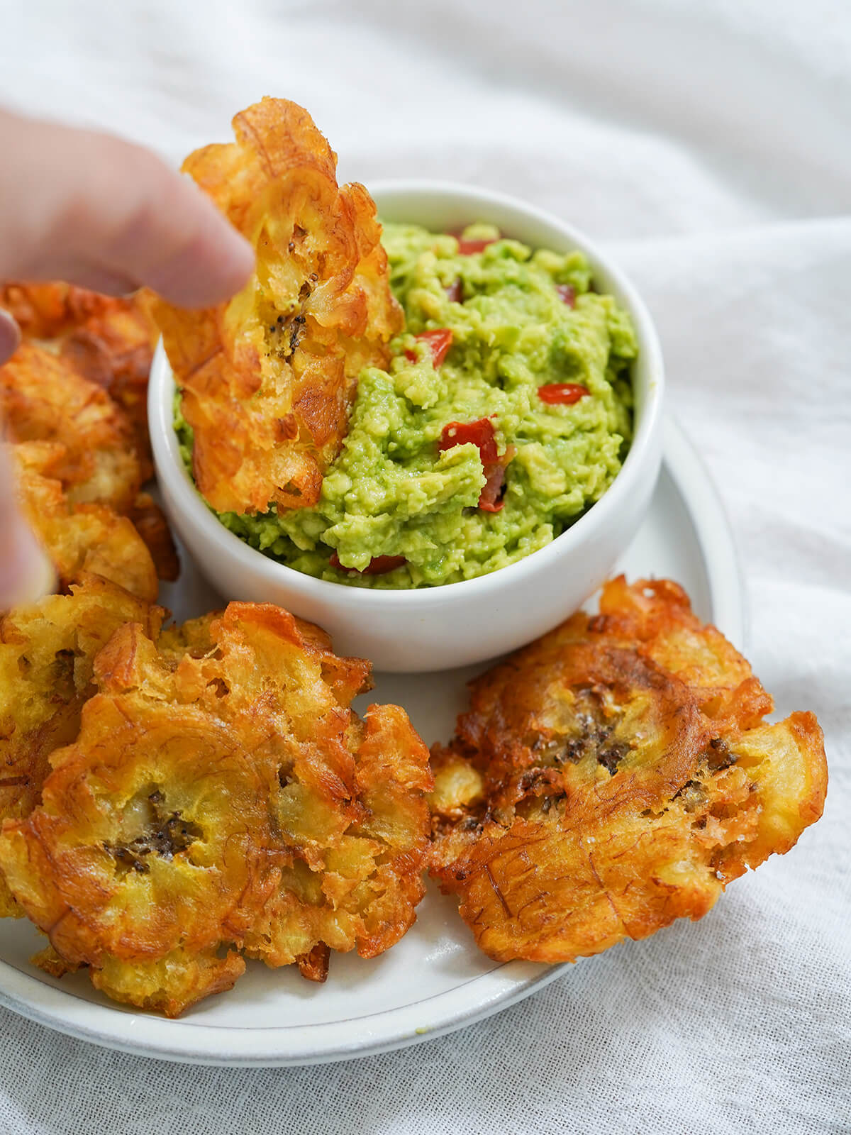 holding a patacone (toston) and dipping it in guacamole with more patacones on plate.