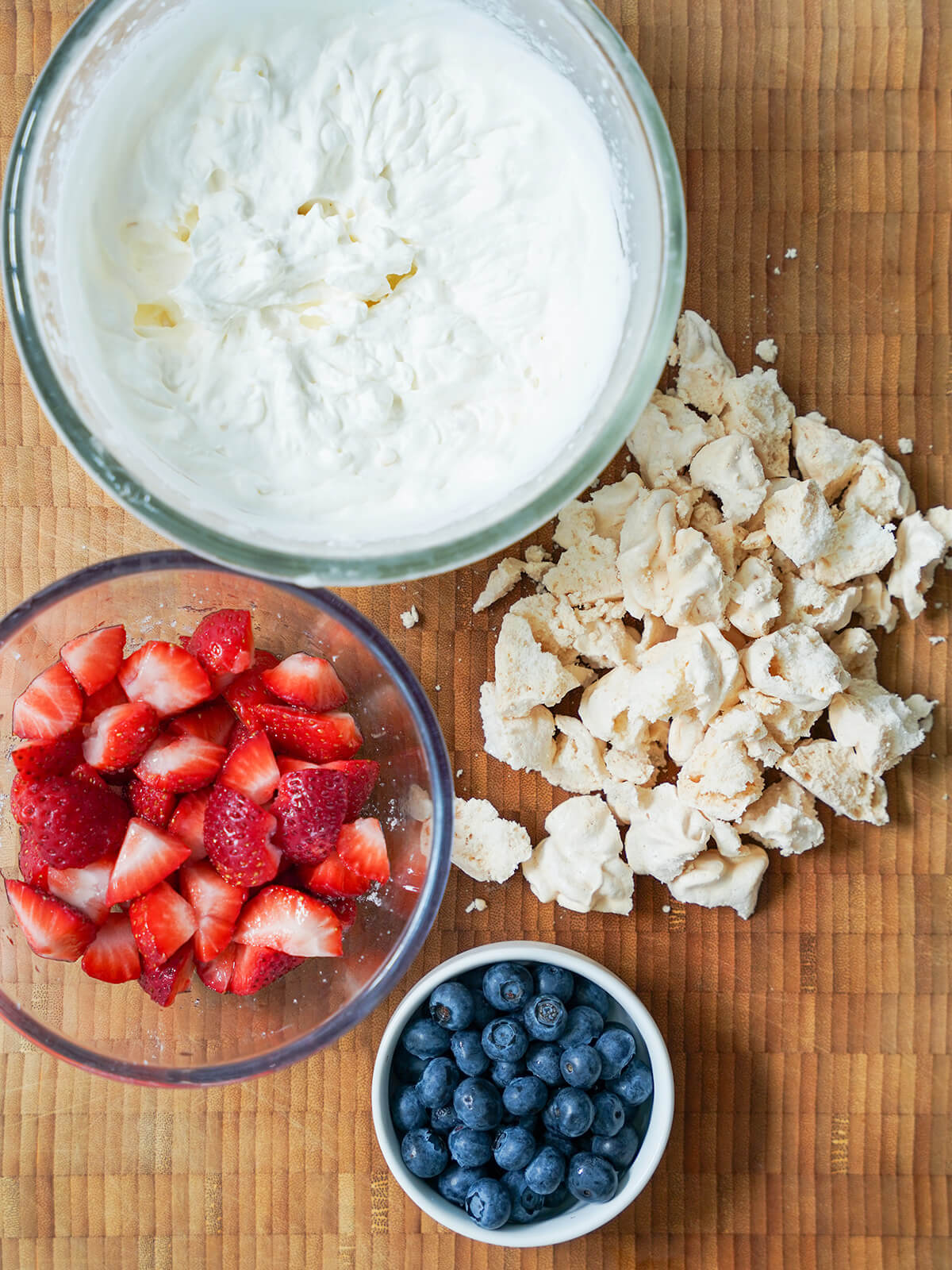 prepared components for Eton mess bowl of whipped cream, bowl of macerated strawberries, bowl of blueberries and broken up pieces of meringue to side.