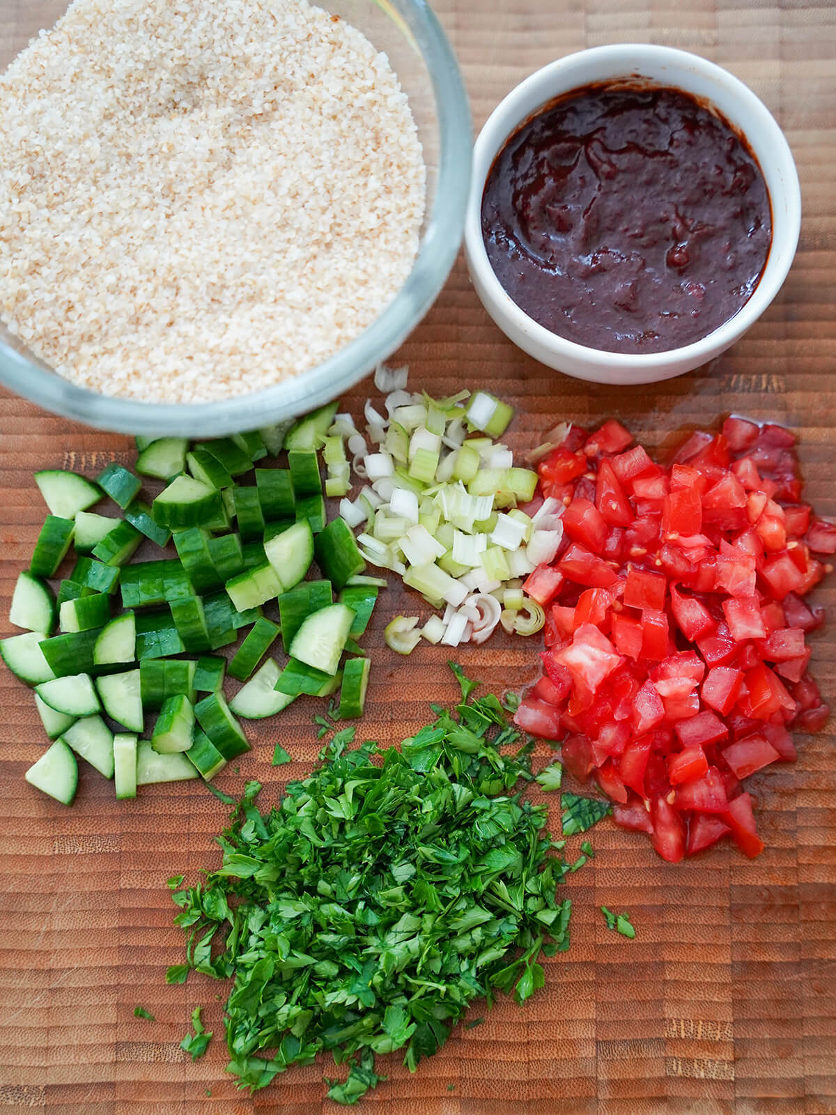 prepared ingredients for kisir bowl of soaked bulgur, small dish with dressing and chopped cucumber, tomato, green onion and parsley on board.