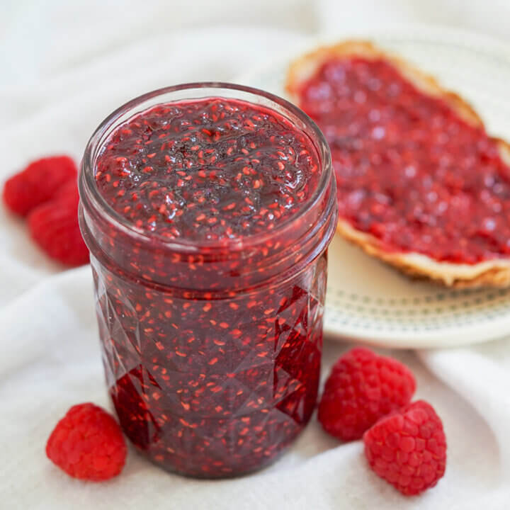 jar of raspberry jam with raspberries either side and blurred toast with jam behind.