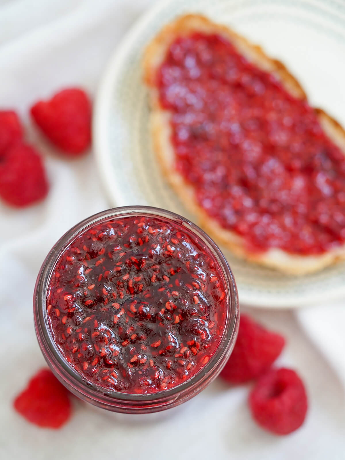 This small batch raspberry jam is easy to make and being lower in sugar, it's packed with bright berry flavor. Delicious on toast, scones, in desserts and more. overhead view of jar of small batch raspberry jam with piece of toast with jam and berries blurred below.