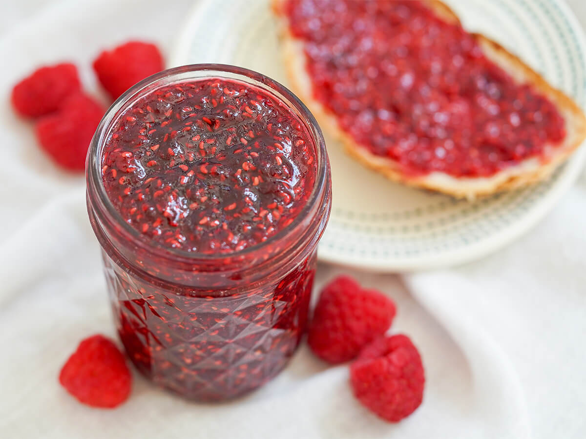 jar of small batch raspberry jam close part overhead view of jar of small batch raspberry jam with raspberries and toast with jam around jar.