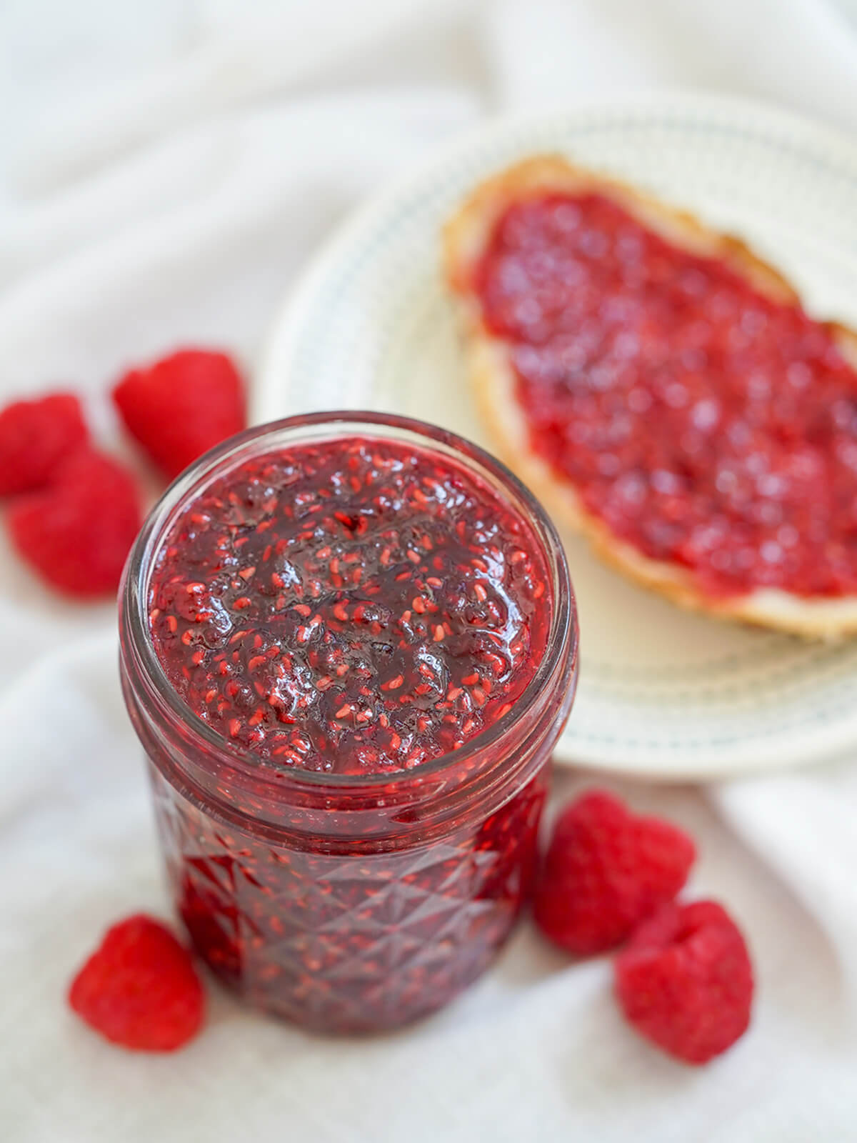 This small batch raspberry jam is easy to make and being lower in sugar, it's packed with bright berry flavor. Delicious on toast, scones, in desserts and more. jar of small batch raspberry jam with blurred piece of toast behind.
