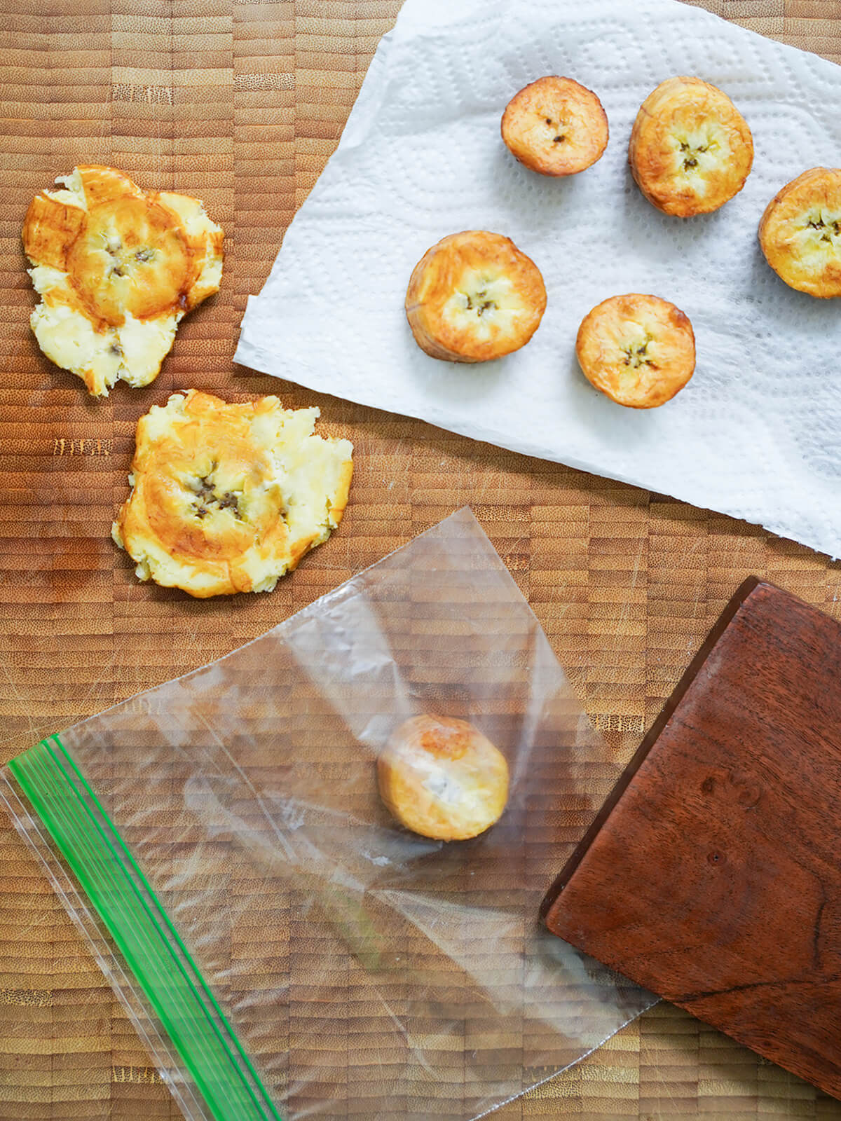 some squashed patacones (tostones) with more fried pieces of plantain draining on paper towel and a slice under plastic with small board for pressing it to side.
