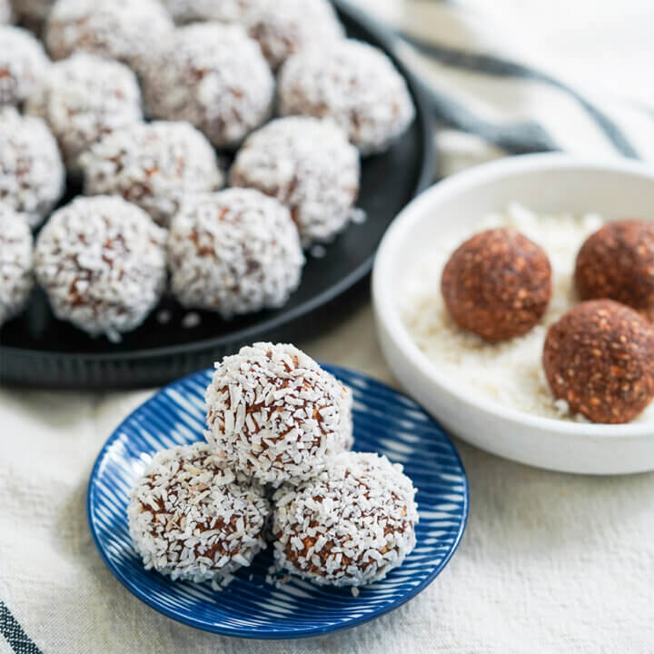 small plate of chocolate oat balls chokladbollar with more behind in background, some finished some still to roll in coconut.