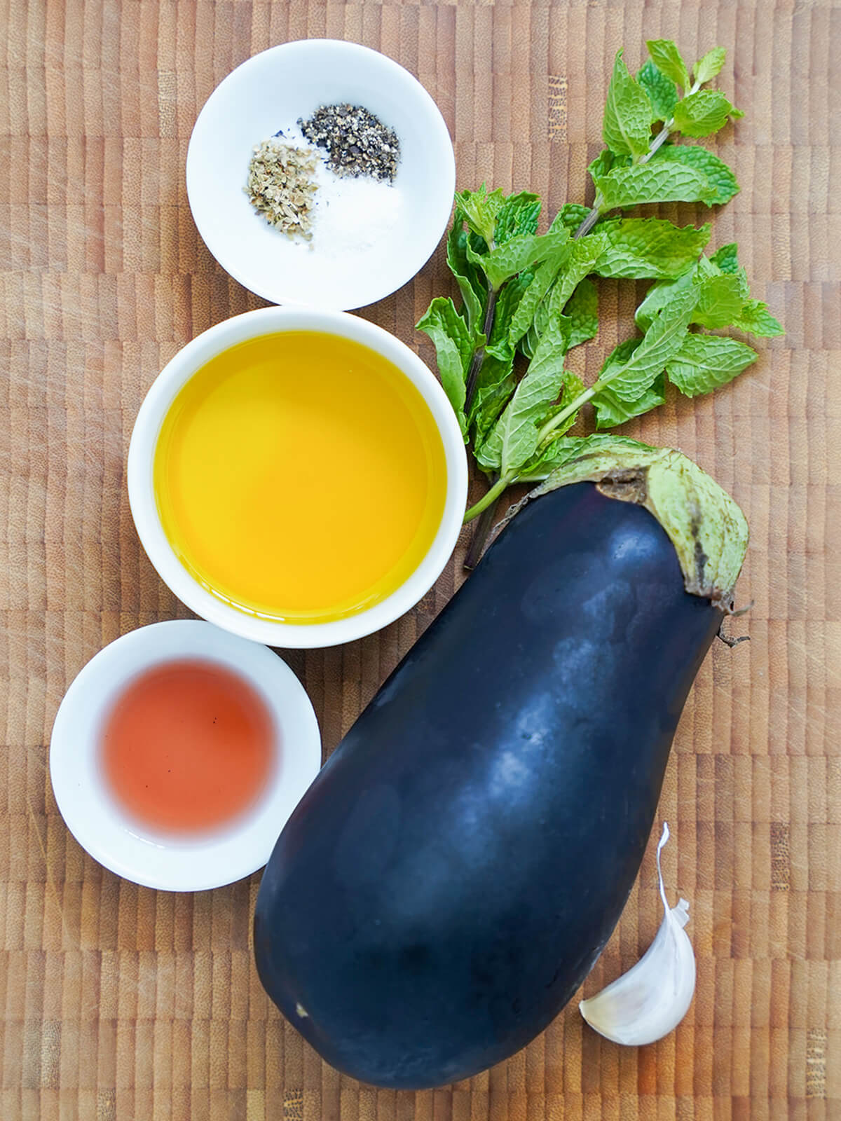 eggplant, bowl of olive oil, small dishes with vinegar, salt and pepper, with sprigs of mint and garlic clove to one side.