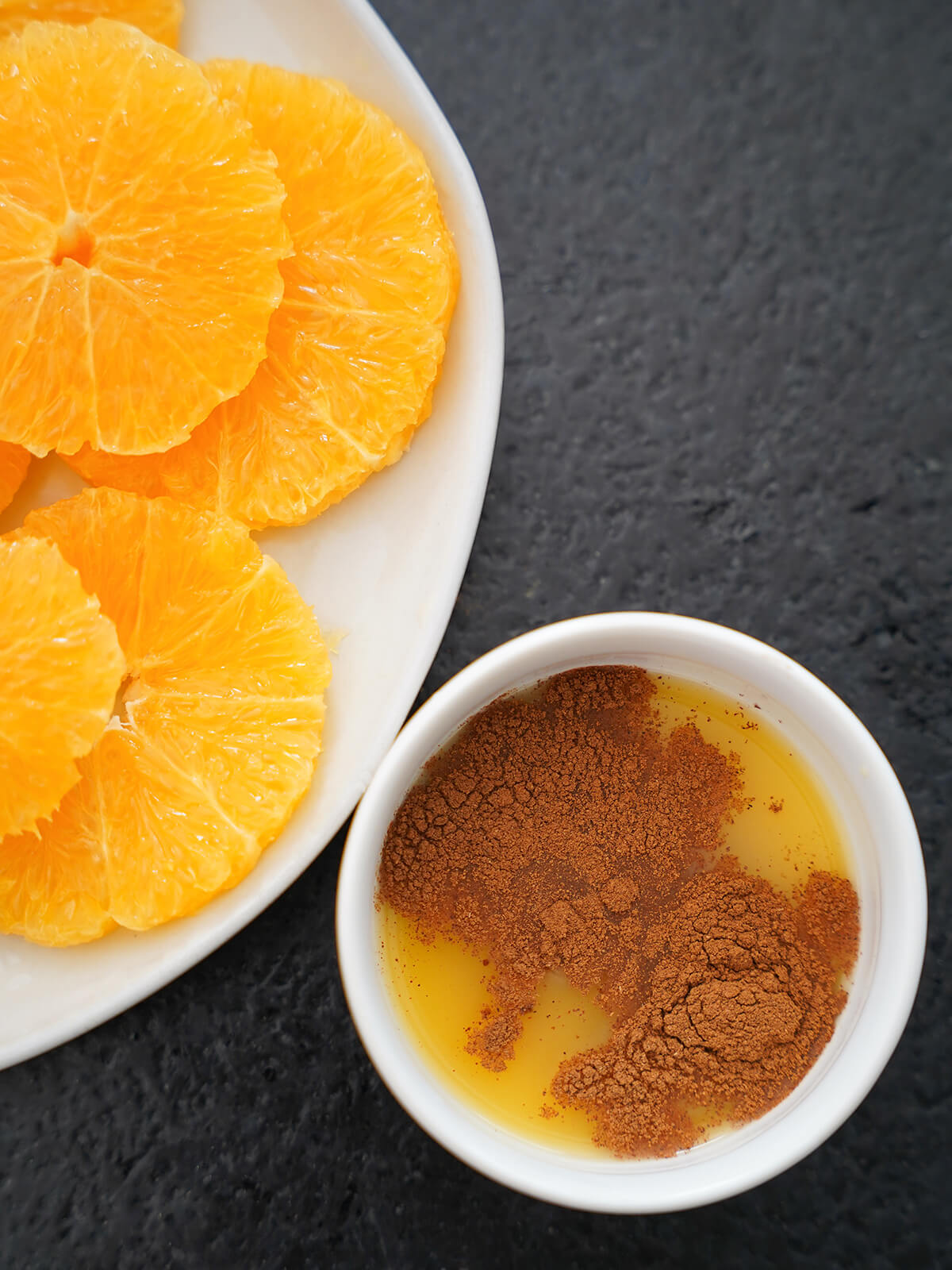 small bowl of orange juice, with cinnamon on top and plate of sliced oranges to side.