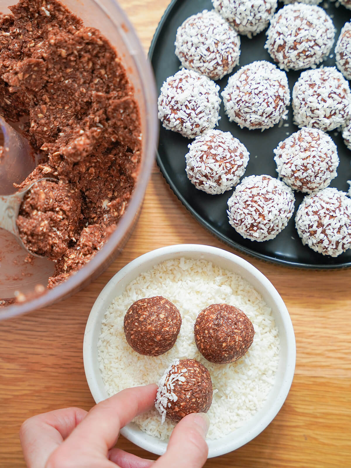 rolling balls of chocolate oat mixture in coconut with some finished to top right and mixture in food processor to left.