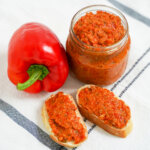 jar of ajvar red pepper spread with some spread on slices of bread in front and pepper to side.