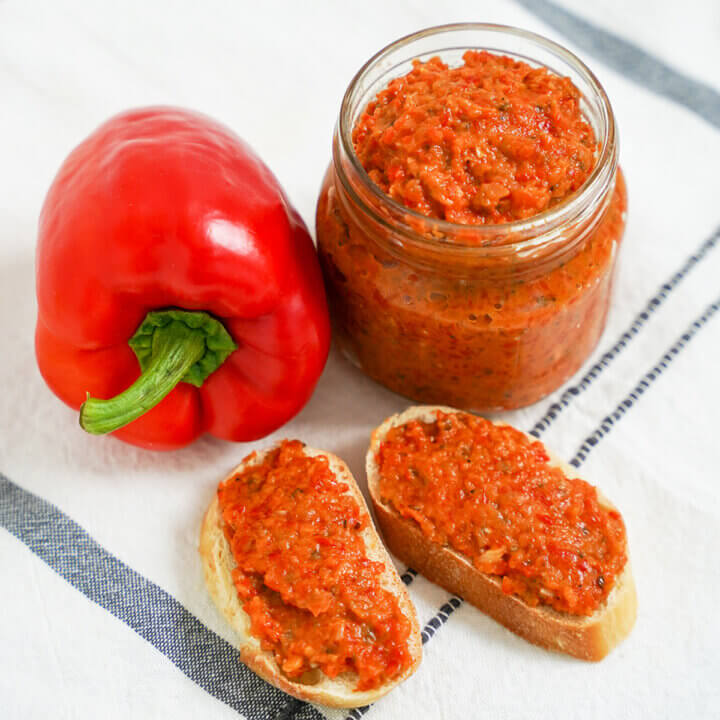 jar of ajvar red pepper spread with some spread on slices of bread in front and pepper to side.