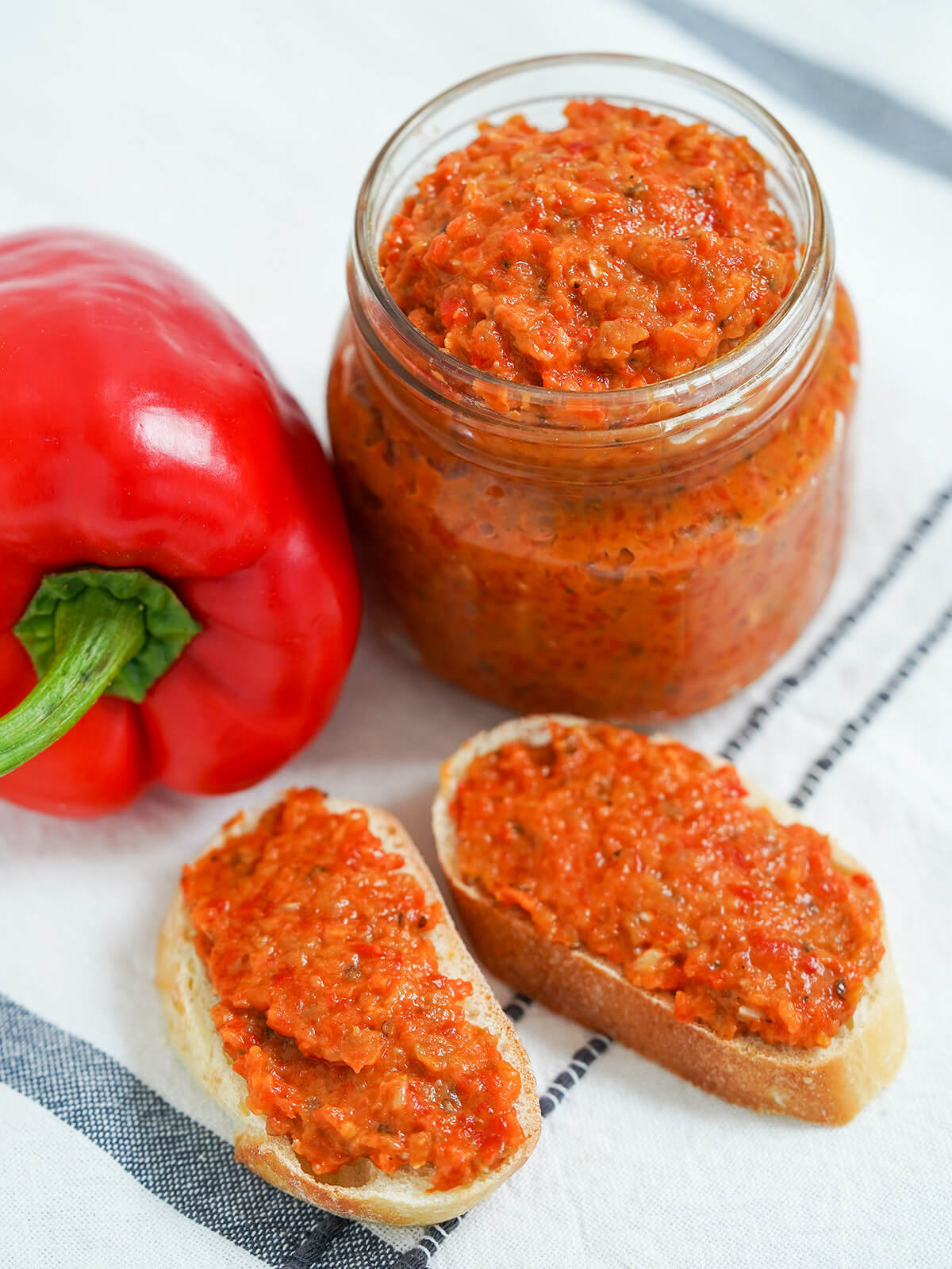 Ajvar is a bright and flavorful Balkan pepper-based dip/spread that's delicious with bread, meats and more. Easy to make and versatile in how you enjoy. jar of ajvar red pepper spread with two slices of bread topped with the spread in front and part view of a red pepper to side.