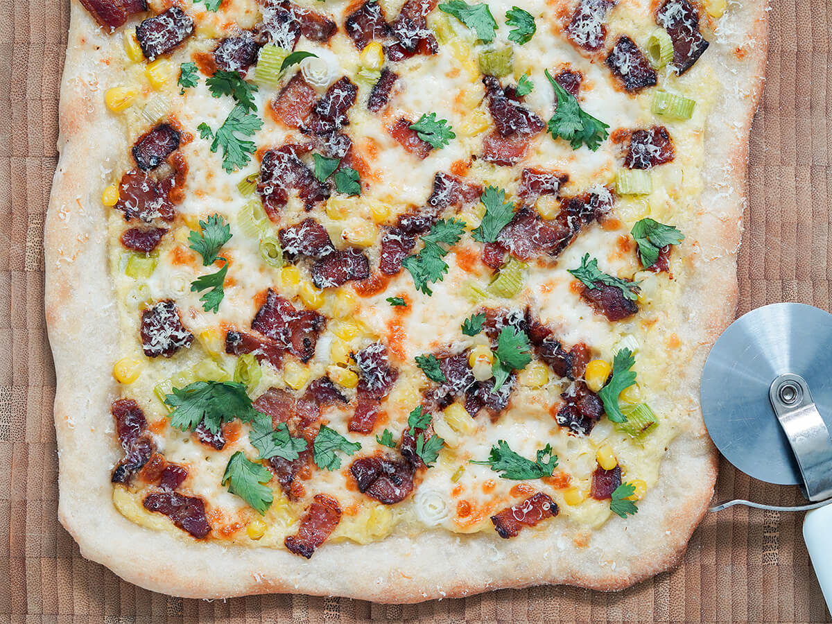 close view of one end of corn pizza with bacon with pizza cutter in corner.