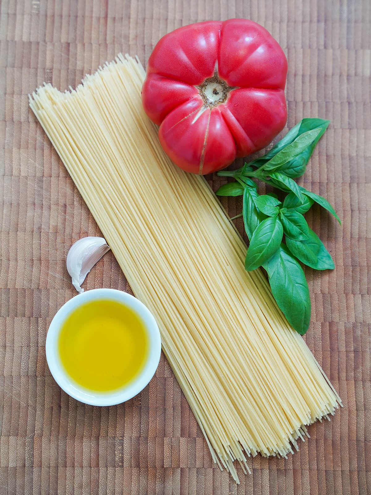 ingredients for pasta with fresh tomatoes stack of spaghetti, large tomato, sprigs of basil, dish with olive oil and clove of garlic on board.