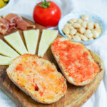 two slices of pan con tomate, Spanish tomato bread, on wooden board with cheese and almonds behind in background.