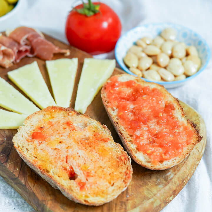 two slices of pan con tomate, Spanish tomato bread, on wooden board with cheese and almonds behind in background.