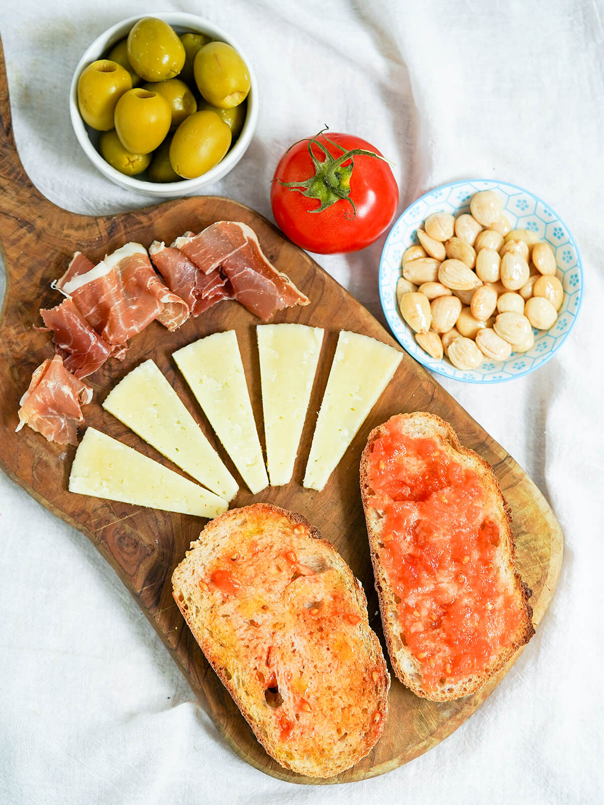 overhead view of wooden board with slices of pan con tomate, Spanish tomato bread, cheese and ham with dishes of almonds and olives to side.