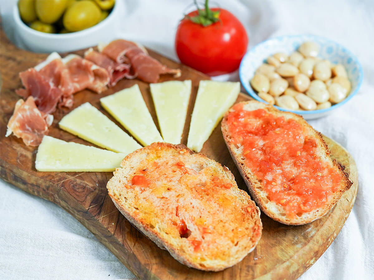slices of pan con tomate, Spanish tomato bread, with slices of cheese and ham on board behind.