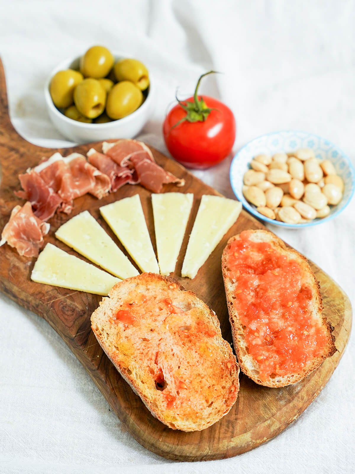 slices of pan con tomate, Spanish tomato bread, on board with cheese and ham slices in rows behind and bowls of olives, nuts and a tomato behind the board.