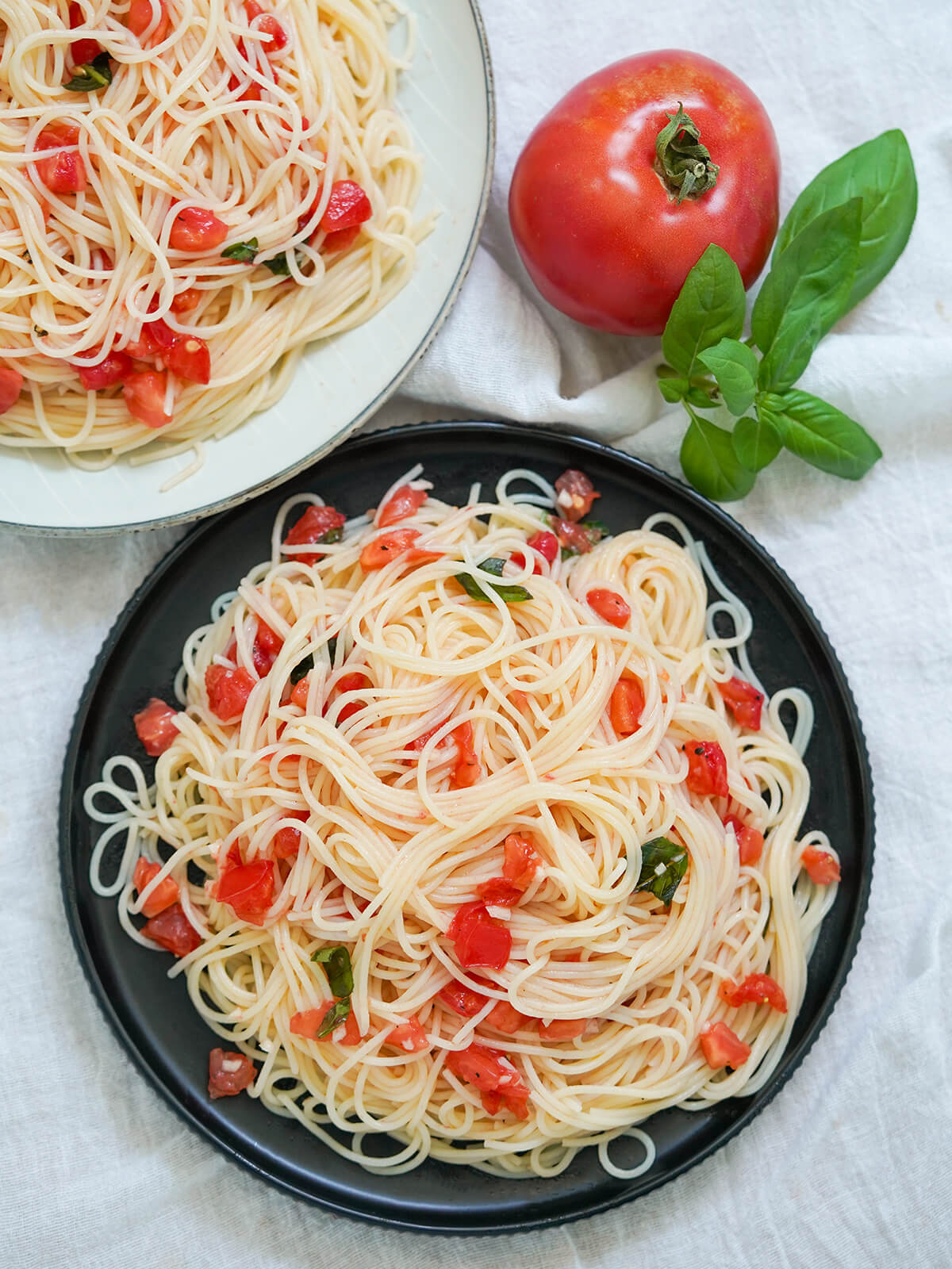 overhead view of a plate of plate of pasta with fresh tomatoes, spaghetti al pomodoro crudo overhead view of plate of plate of pasta with fresh tomatoes, spaghetti al pomodoro crudo with second plate in corner and tomato to one side.