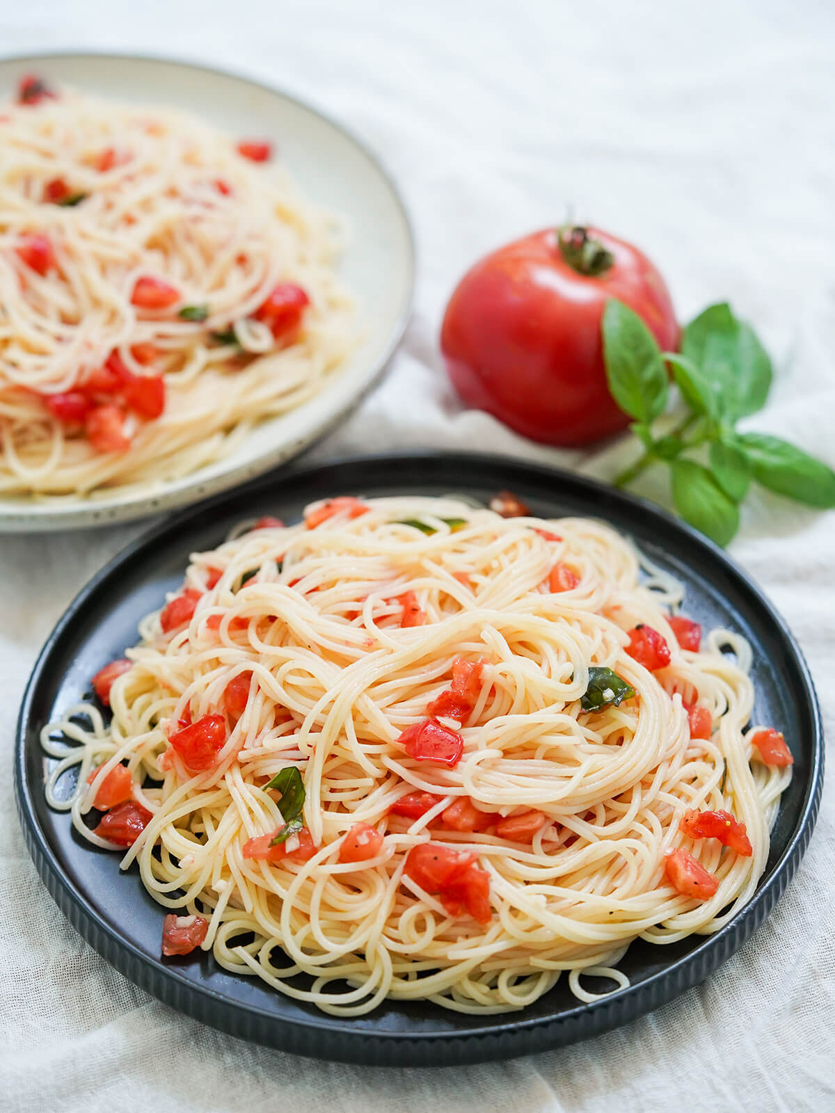 Pasta with fresh tomatoes (spaghetti al pomodoro crudo) is easy to make with a simple no cook sauce that's perfect for summer tomatoes. Quick, bright and so tasty. plate of plate of pasta with fresh tomatoes, spaghetti al pomodoro crudo with tomato and sprig of basil behind and part of another plate in background.