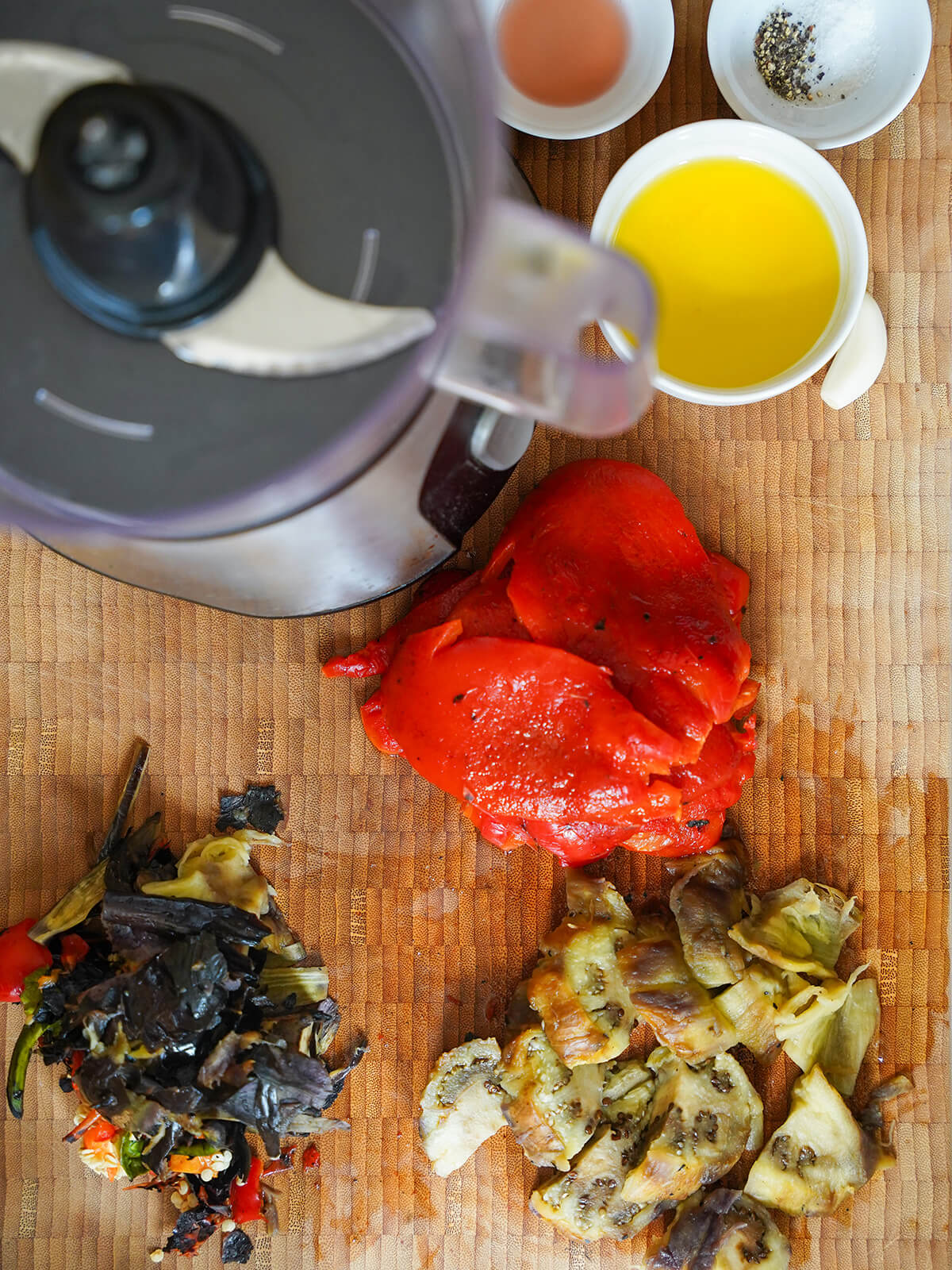 preparing ingredients for ajvar red pepper spread overhead view of peeled charred pepper and eggplant pieces with dishes of oil, vinegar and a garlic clove to side next to food processor.
