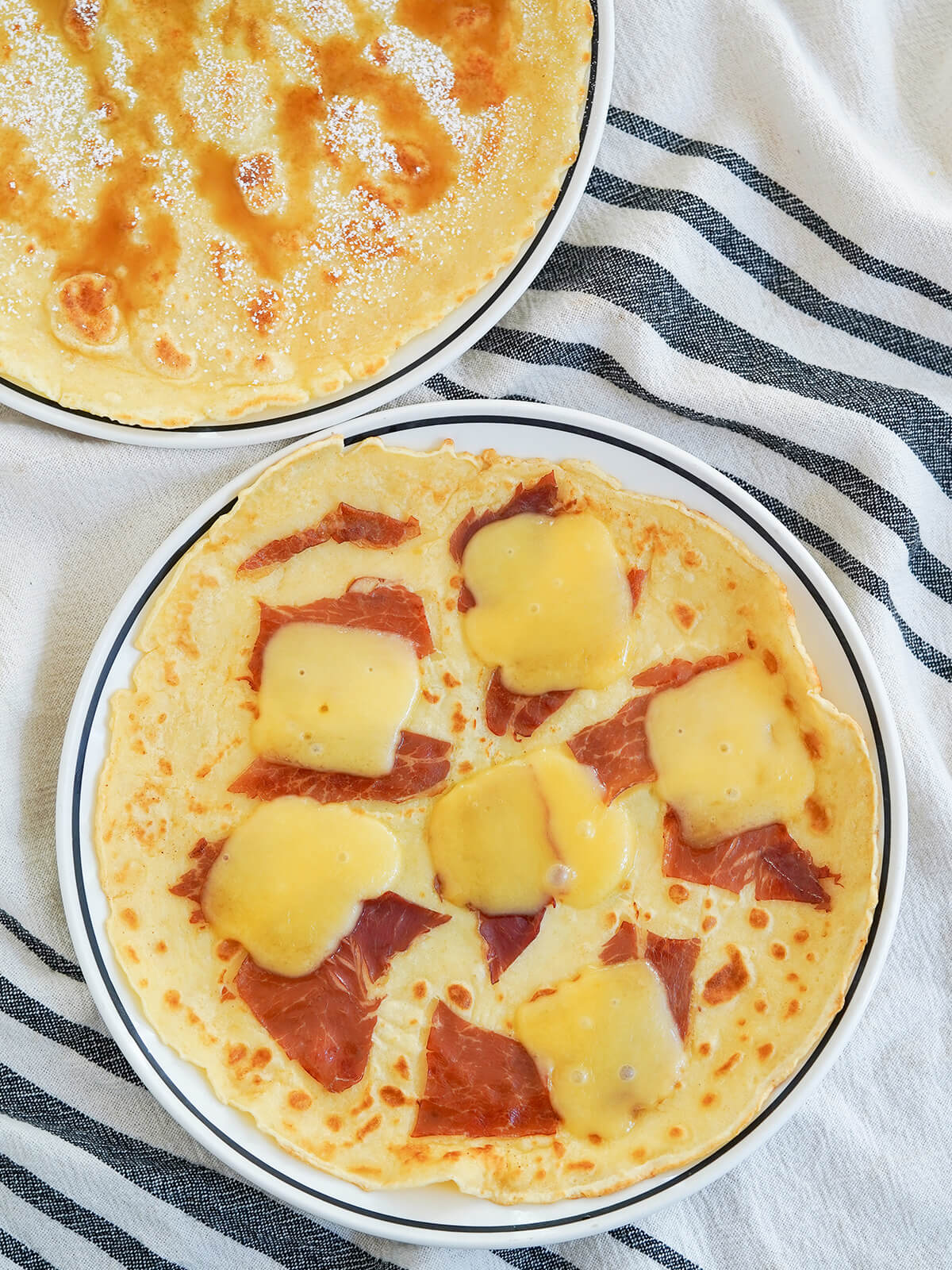 overhead view of two plates of Pannekoeken Dutch pancakes with one topped with ham and cheese and other syrup and powdered sugar.