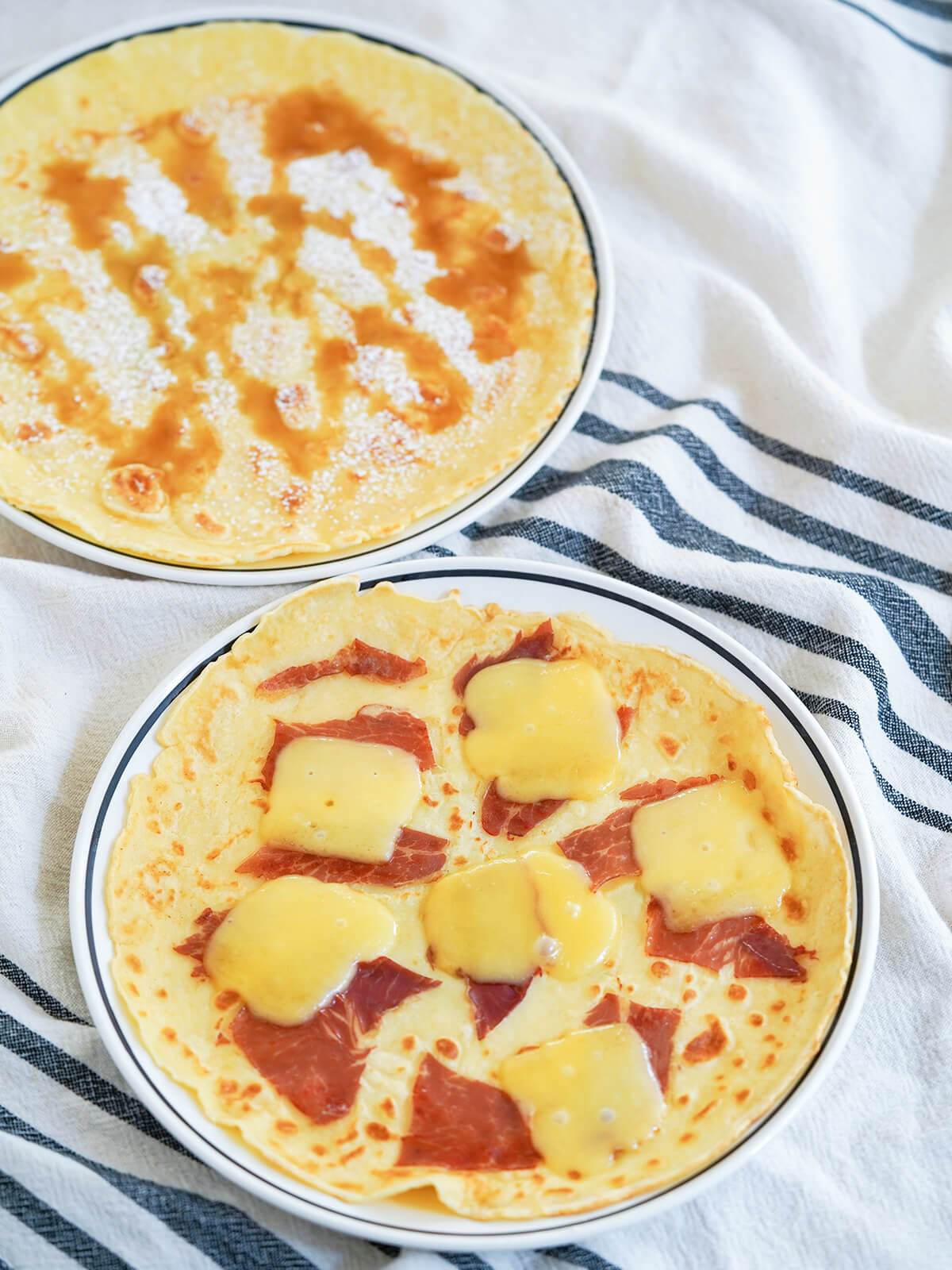 Dutch pancakes, pannekoeken, are wide and thin, and a blank canvas for both sweet and savory toppings. Comforting, adaptable and sure to be a favorite. plates of Pannekoeken Dutch pancakes, front with ham and cheese and behind with syrup and sugar.