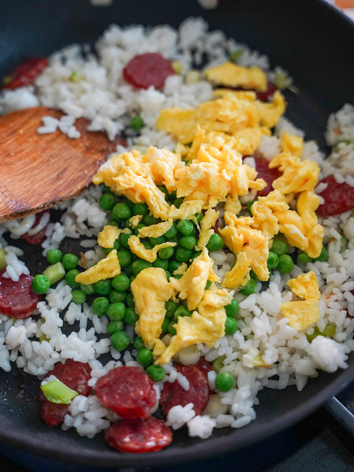cooking Taiwanese fried rice rice, slices of sausage in pan with peas and egg pieces on top and spatula to side.