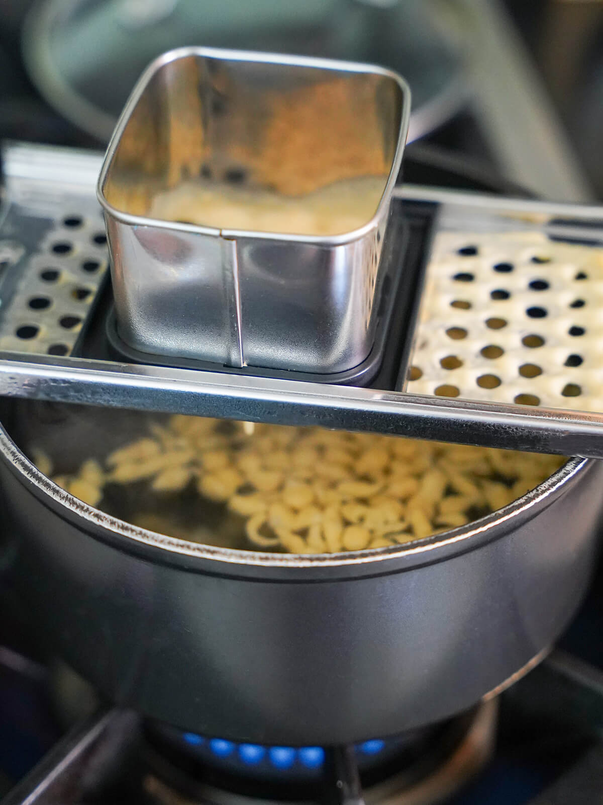 cooking spaetzle by pressing batter through spaetzle maker. speatzle maker over pot of water with some spaetzle floating in water in pot.