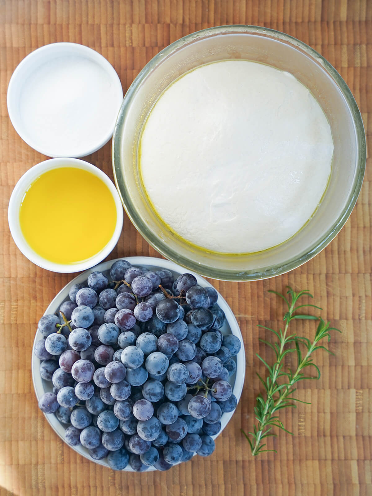 bowl of bread dough, small dishes with sugar and olive oil, plate of grapes and sprigs of rosemary on wooden board.