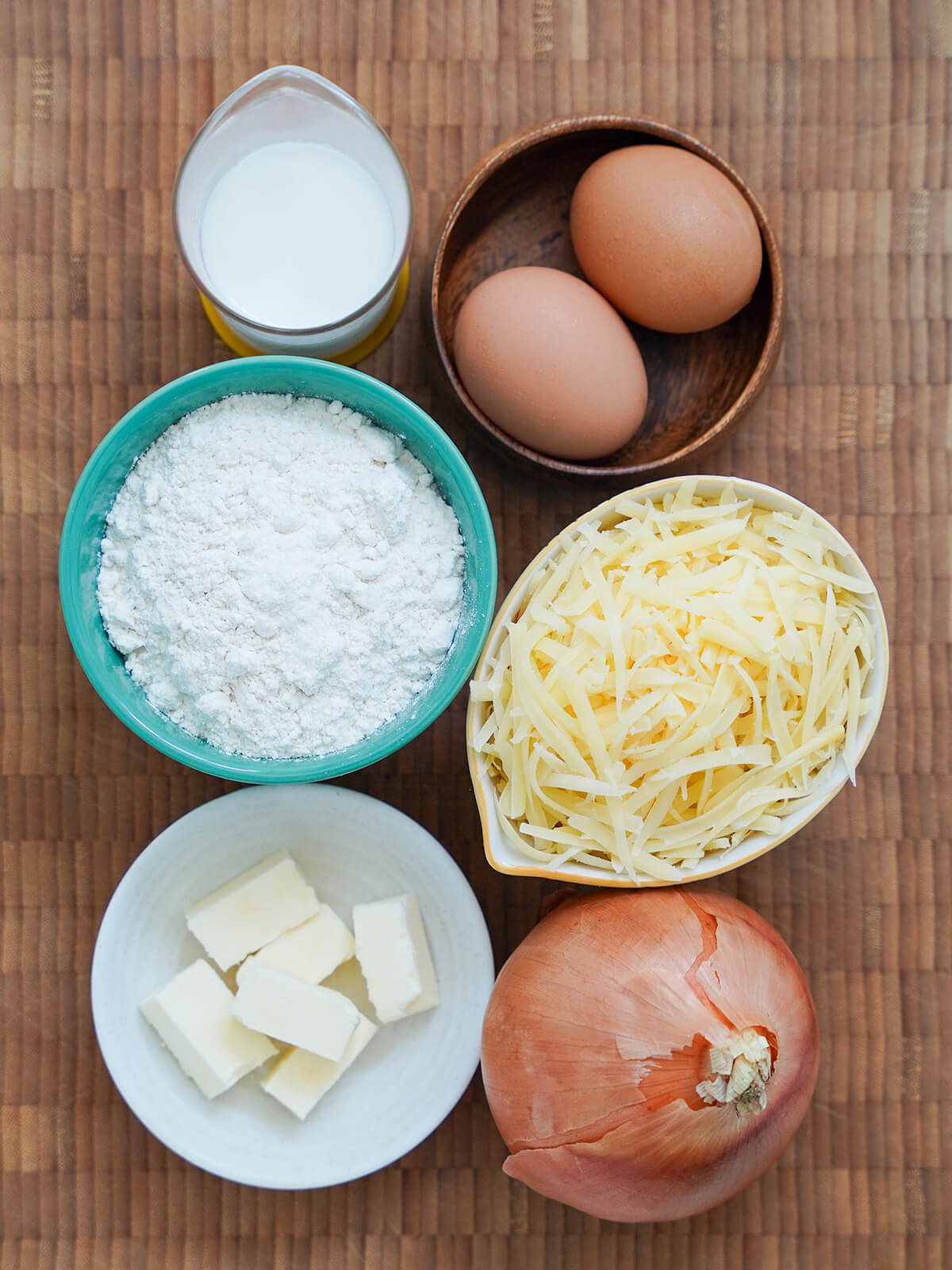 ingredients for Käsespätzle bowls with eggs, flour, grated cheese, butter, measure of milk and onion below on wooden board.