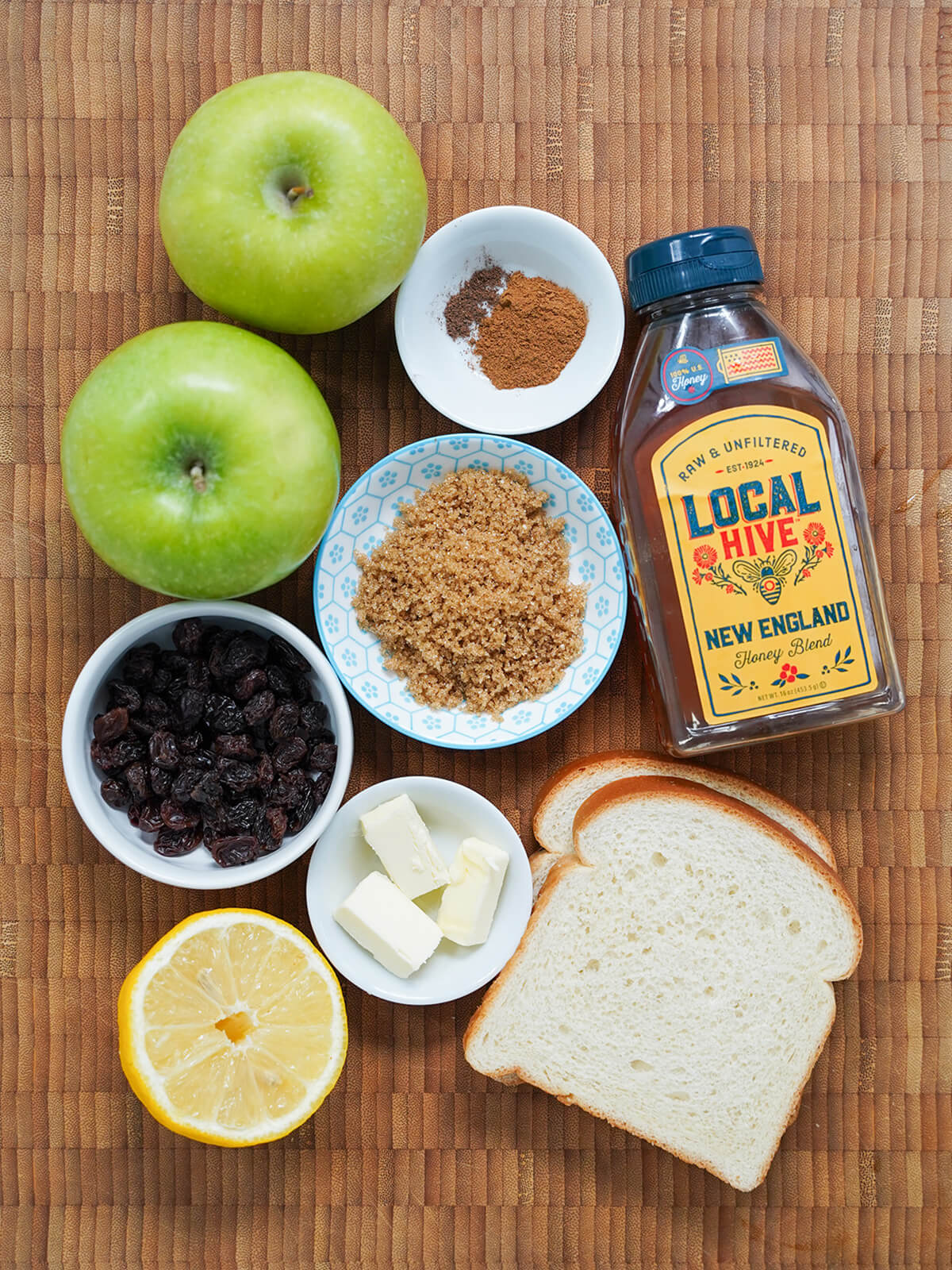 apples, dishes of sugar, raisins spices. slices of bread, part lemon, bottle of honey on wooden board.