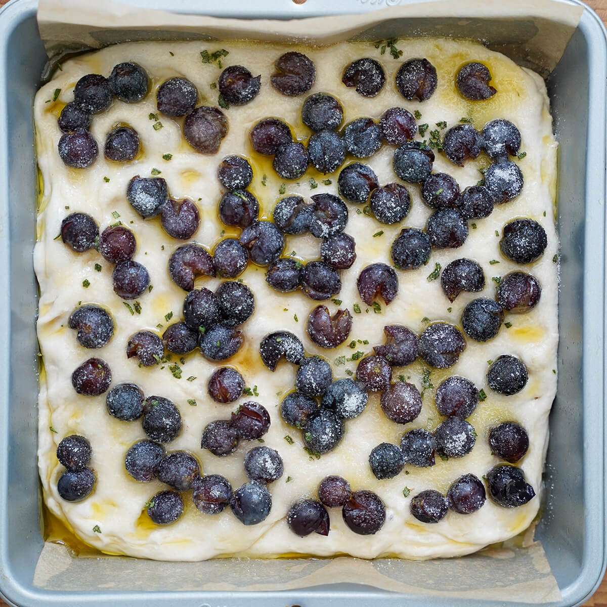 bread dough in baking pan topped with grapes, chopped rosemary, sugar and olive oil before baking.