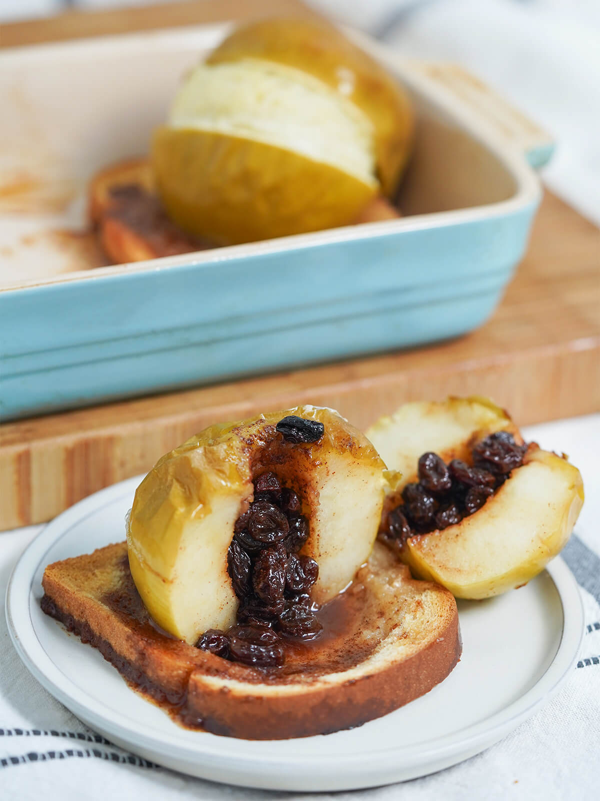 baked apple cut open showing raisin filing and cinnamon mixture running onto bread with another stuffed apple in dish behind.