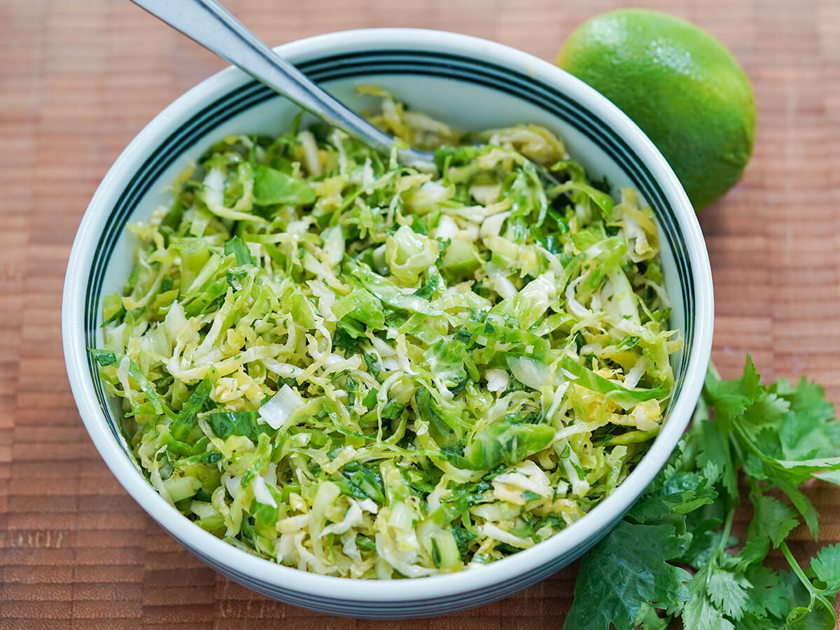 bowl of Brussels sprouts slaw with stems of cilantro under bowl and lime behind.