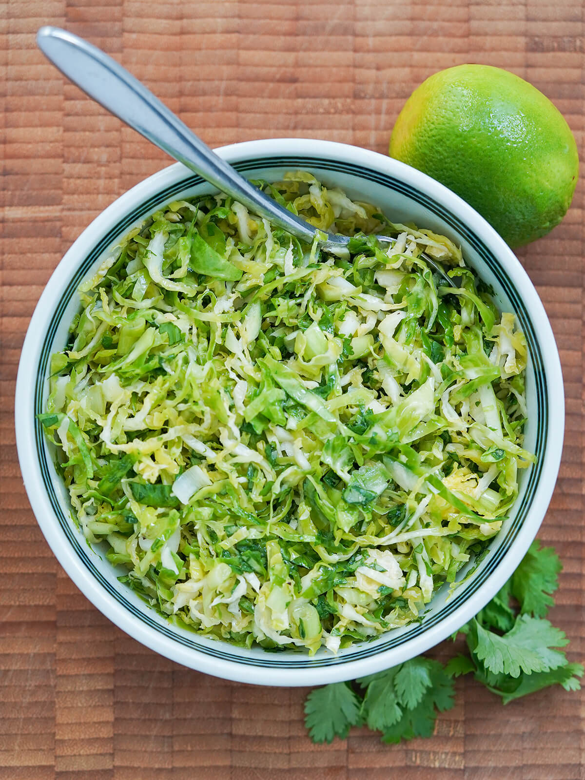 overhead view of bowl of Brussels sprouts slaw with spoon in bowl, lime above and cilantro below.