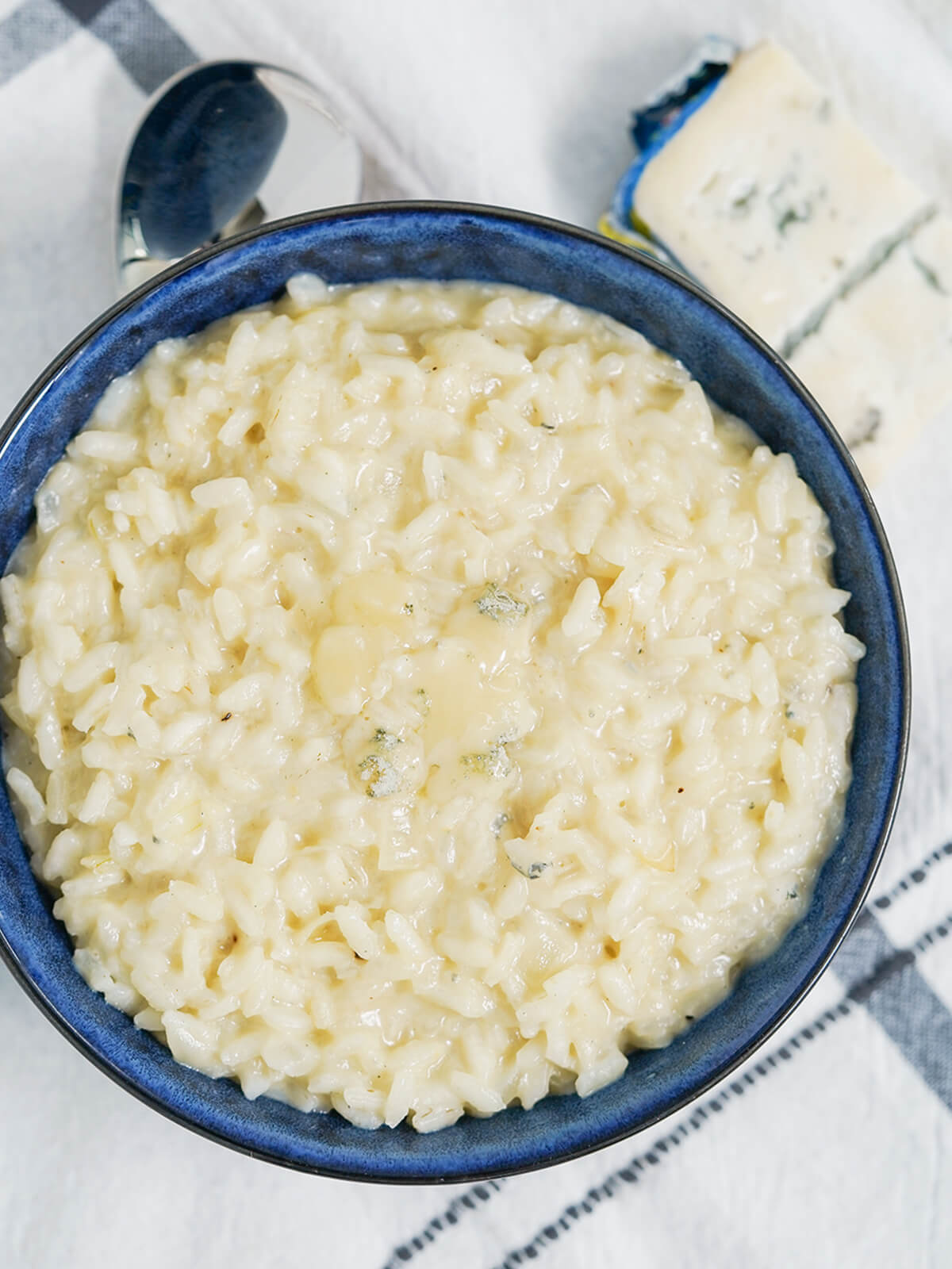 This gorgonzola risotto is easy to make and full of fantastic flavor. Creamy and comforting, it's great on it's own or served as a side. overhead view of bowl of gorgonzola risotto with spoon under bowl and part block of cheese in top right corner.