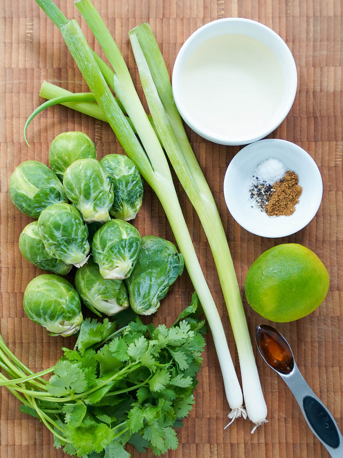 Brussels sprouts, green onions, cilantro, bowls with oil, cumin, salt and pepper, lime and measure of honey on wooden board.