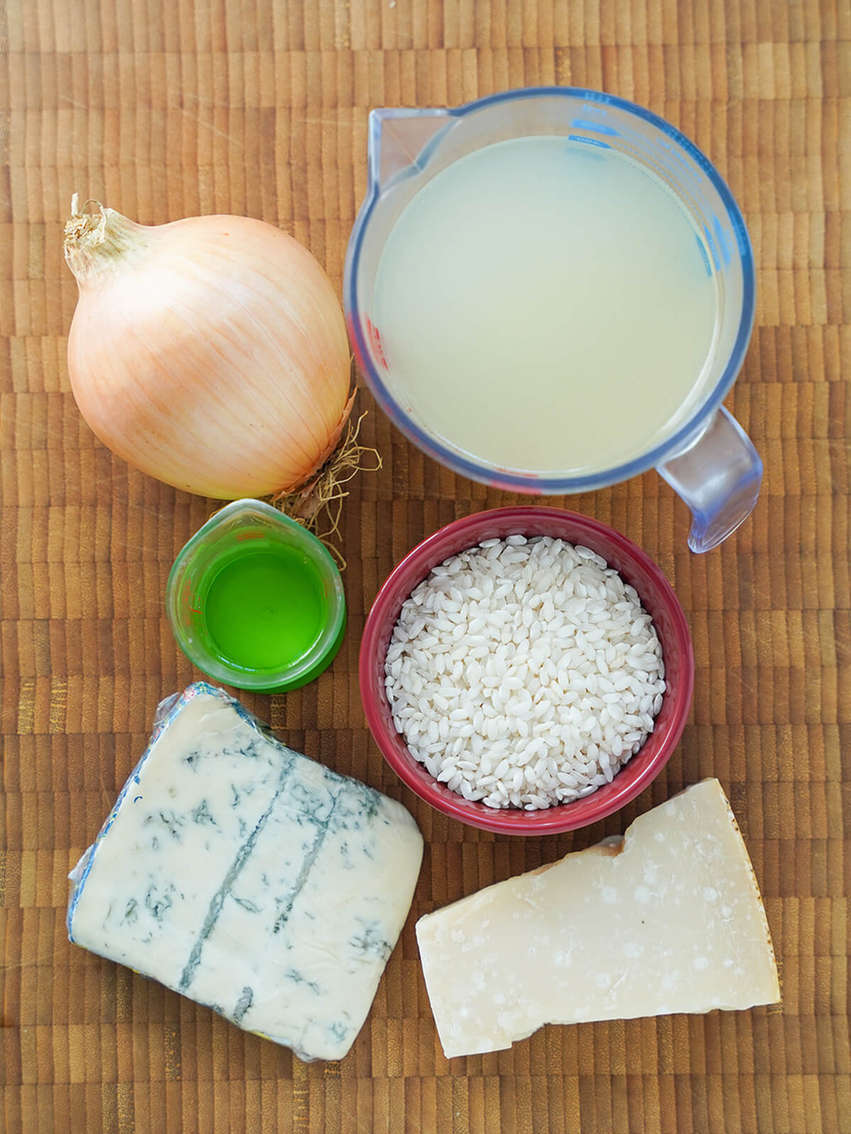 ingredients for gorgonzola risotto measure of stock, onion, bowl of rice, blocks of parmesan and gorgonzola cheese and small measure of white wine on wooden board.