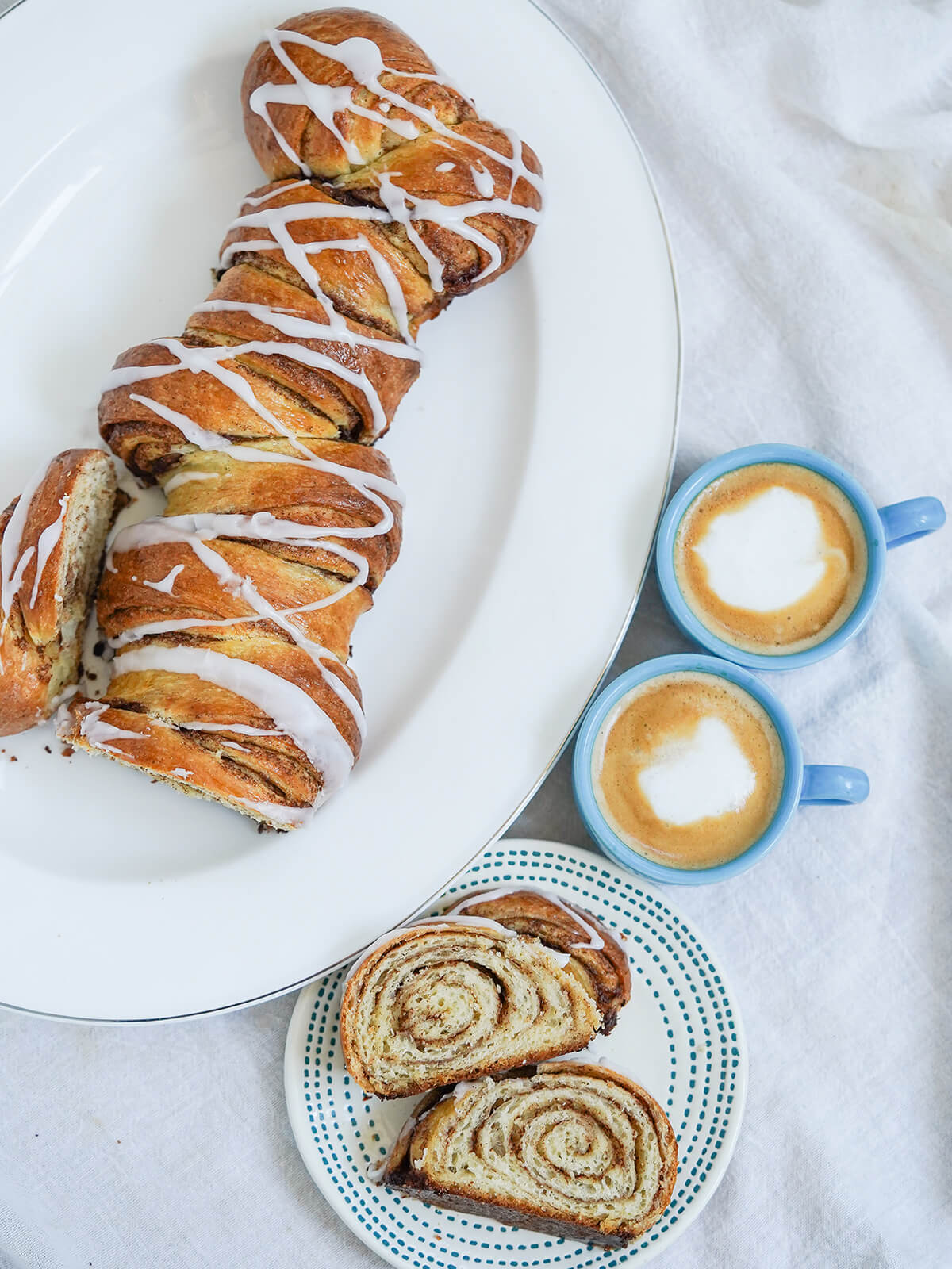 This kanelstang, Danish cinnamon twist, is a bit like a cinnamon roll loaf, a delicious mix of buttery, soft, cinnamon-filled and gently crisp on the outside. A perfect coffee time treat. overhead view of most of a kanelstang, Danish cinnamon twist with two slices on a plate below and cups of coffee to side.