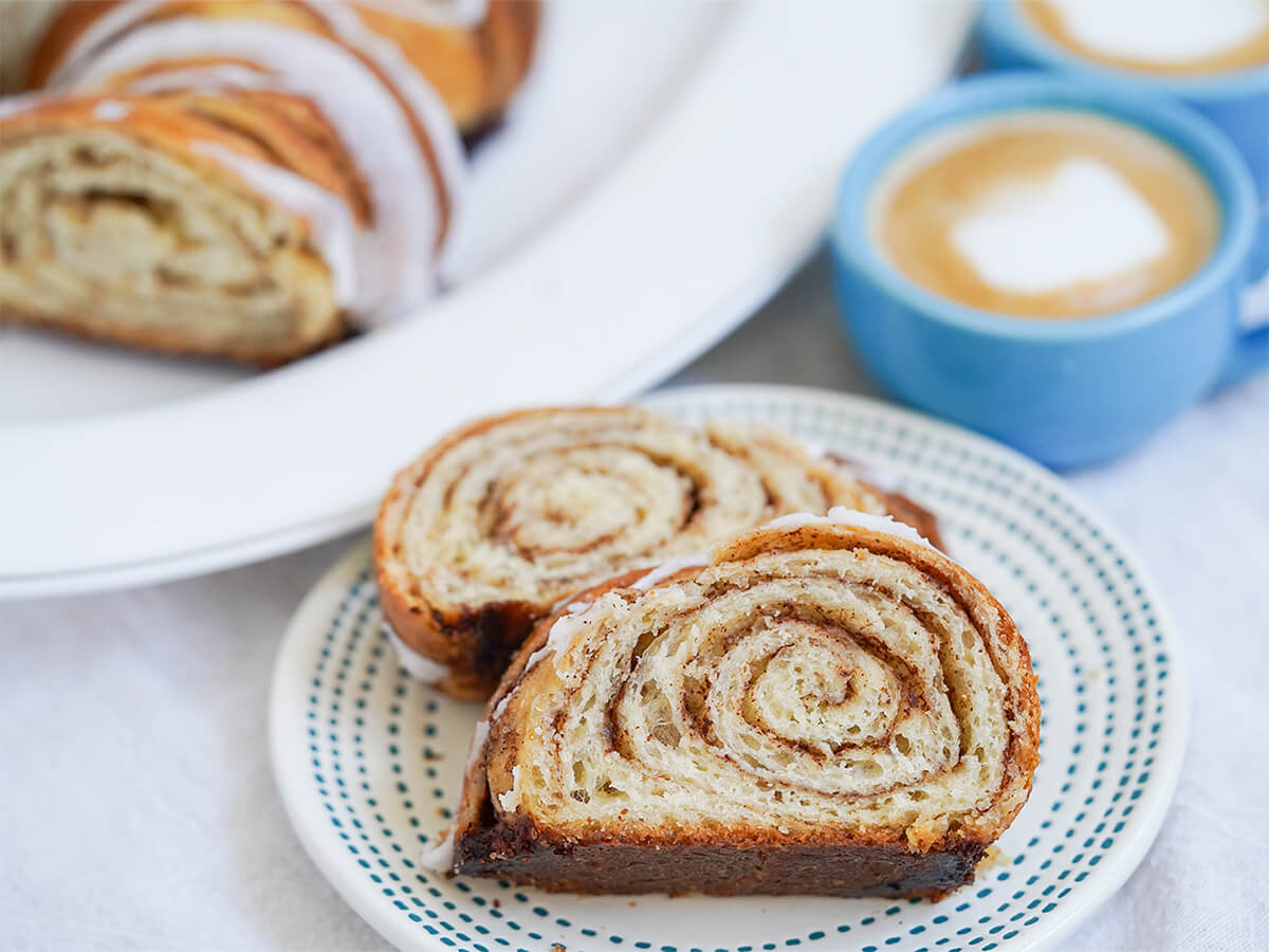 kanelstang Danish cinnamon twist two slices of kanelstang, Danish cinnamon twist, on a plate with more and cups of coffee blurred in background behind.