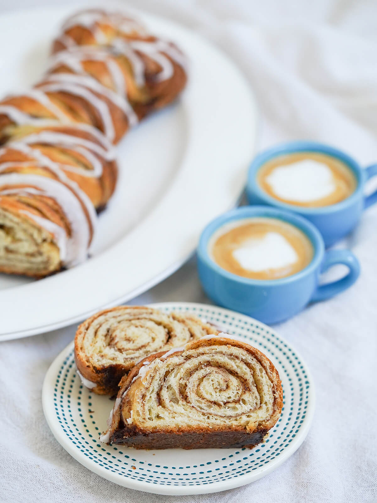 This kanelstang, Danish cinnamon twist, is a bit like a cinnamon roll loaf, a delicious mix of buttery, soft, cinnamon-filled and gently crisp on the outside. A perfect coffee time treat. two slices of kanelstang Danish cinnamon twist on small plate with rest of loaf behind and two cups of coffee to side behind plate.